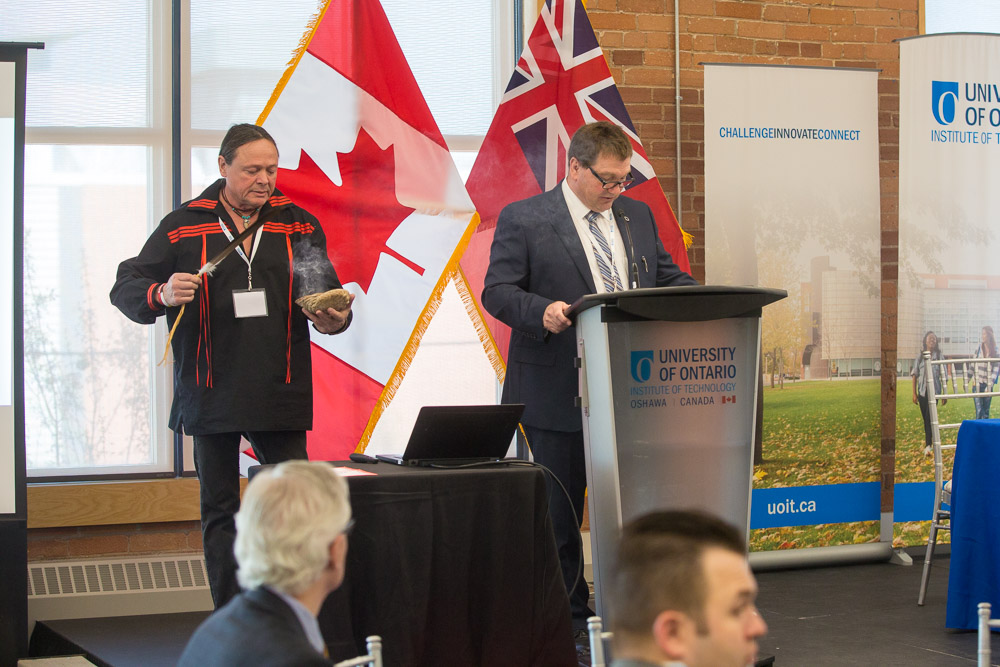 Rick Bourque, Traditional Knowledge Keeper, UOIT-Baagwating Indigenous Student Centre (left), performs traditional smudging ceremony prior to the start of the 2017 Futures Forum (at podium: emcee Doug Holdway, PhD, Interim Vice-President, Research, Innovation and International).