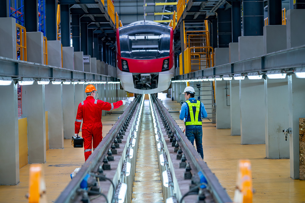 Two professional workers stand in front of an electric train inside a facility, viewed from behind and slightly below. The workers are observing the train, which is elevated on tracks for construction or maintenance. The sleek design of the train contrasts with the industrial setting of the facility.