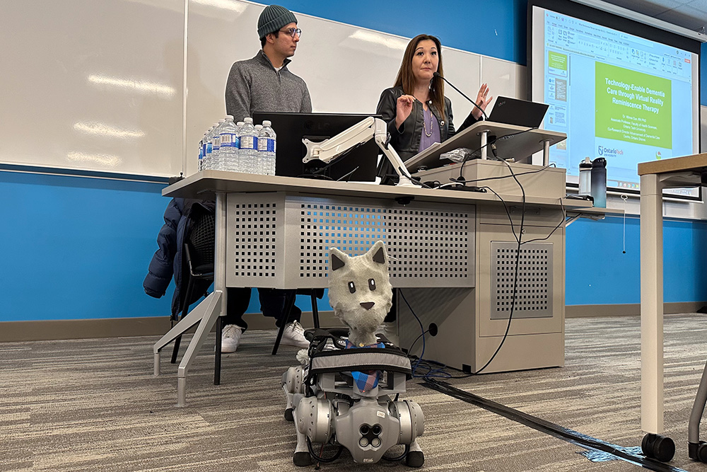 Presenting 'Brando', a robotic dog able to express emotion with the use of a robotic tail and ears. Ontario Tech University researcher Dr. Winnie Sun of the Faculty of Health Sciences (top right) and graduate student Marco Martinez discuss social robotics and virtual reality reminiscence therapy at the Ontario Tech/Lakeridge Health PATH Research Connection Day (Software and Informatics Research Centre, December 3, 2024).