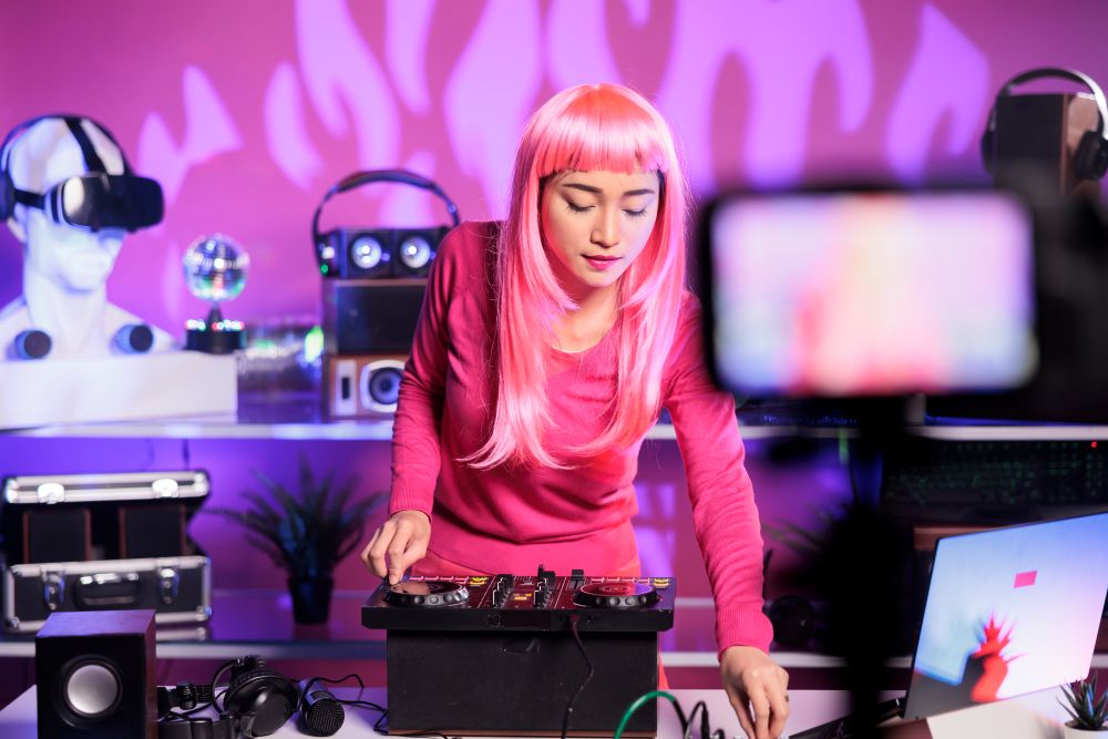 Young woman with straight, pink hair is standing in front of a DJ setup. In the background, partially obscured by a cell phone camera, are a mannequin head wearing VR glasses, a disco ball, speakers and a laptop. 